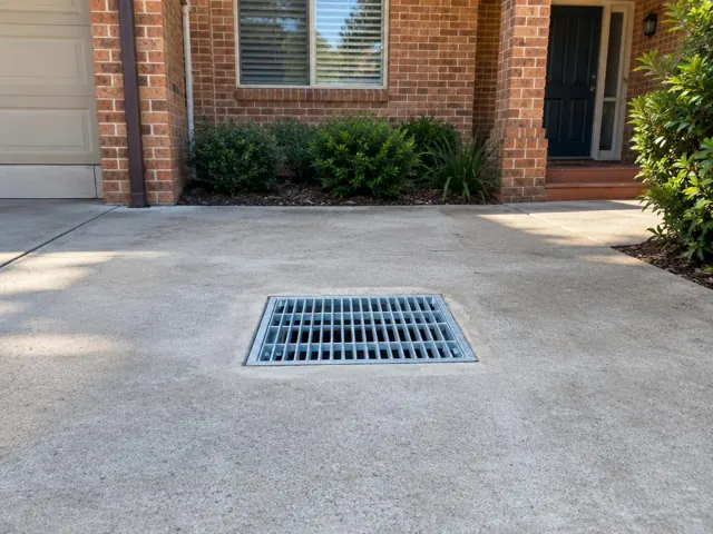 Concrete driveway with a clean metal drain grate near a brick house and front entry in Pennant Hills